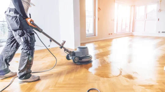 Un spécialiste sable un plancher de bois franc dans une salle de séjour résidentielle, la bande de bois brut fraîchement poncé contrastant avec l'ancien vernis.